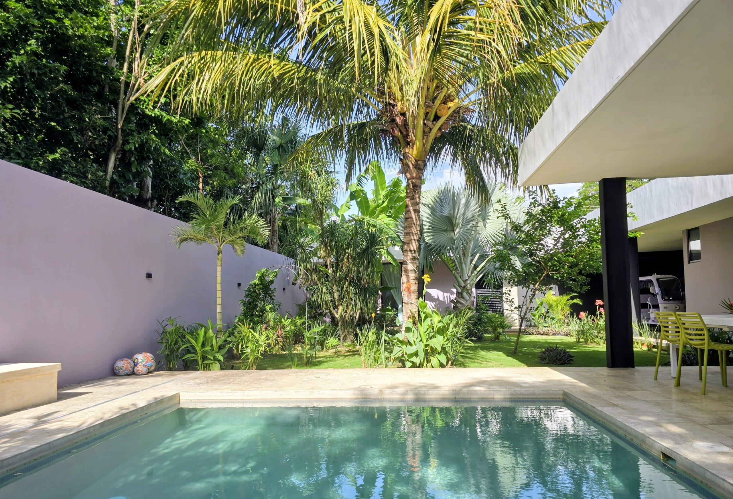 A sunlit swimming pool reflects a tall palm and lush tropical garden beside a purple wall and modern covered patio.